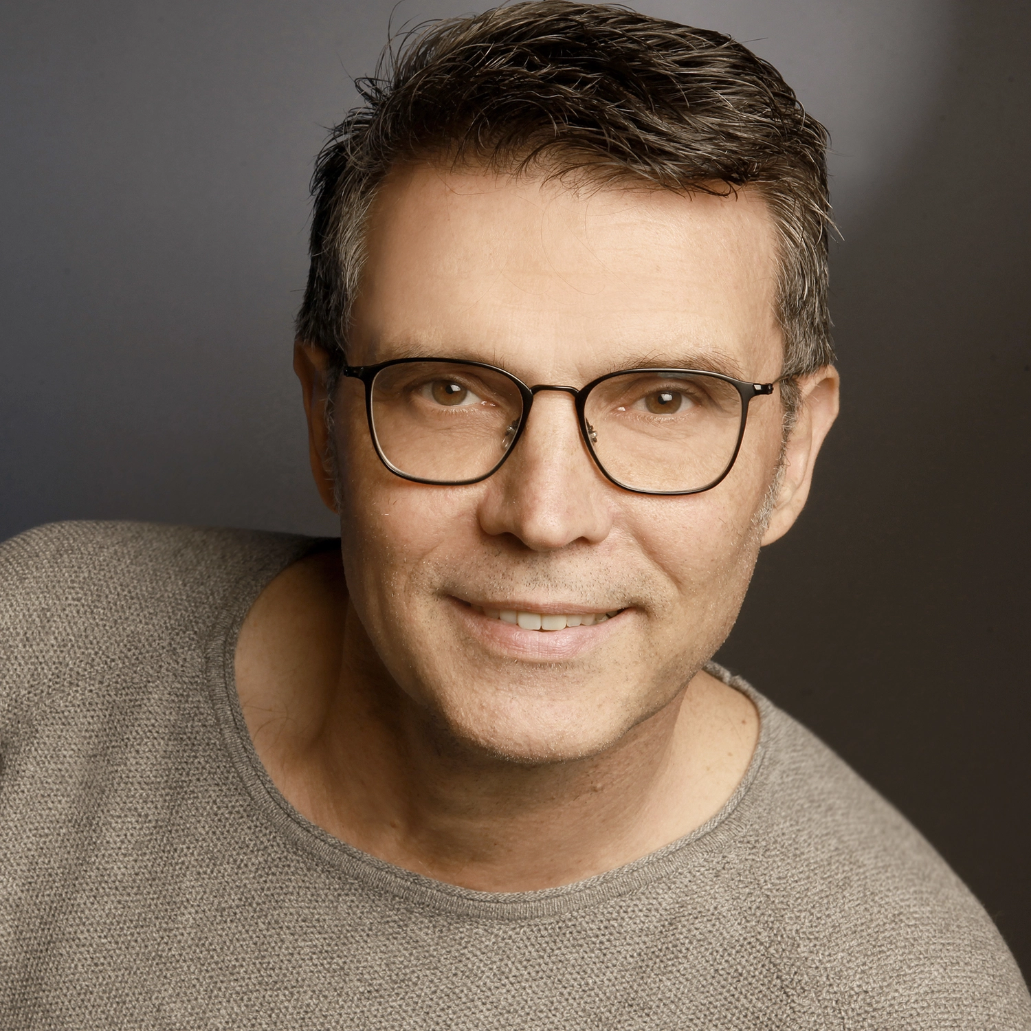 Dein Coach Kai – Executive & Leadership Coaching – Nahaufnahme eines lächelnden Mannes mittleren Alters mit Brille, der ein gemustertes blaues Button-Down-Hemd trägt, vor einem dunkelgrauen Hintergrund.